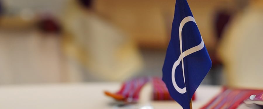 Miniature Indigenous infinity loop flag with a red Metis sash in the foreground and traditional beadwork design overlay